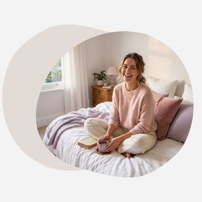 jeune femme souriante et détendue assise sur un lit aux draps de lin blanc, lumière naturelle chaleureuse de fin de journée