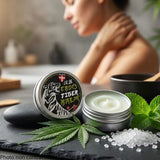  In the foreground, a small, round, vintage-style metal tin sits open on a smooth, dark slate stone, revealing a thick, white-mint-hued balm. Beside the tin, fresh green hemp leaves, a sprig of refreshing peppermint, and tiny white camphor crystals visually evoke the potency of the botanical ingredients.
