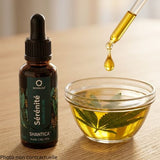 The bottle rests on a light oak table with a natural, minimalist look. Beside it, fresh green hemp leaves are gently infusing in a small vintage glass bowl filled with golden vegetable oil, illustrating the full-spectrum natural maceration extraction method (no powders)